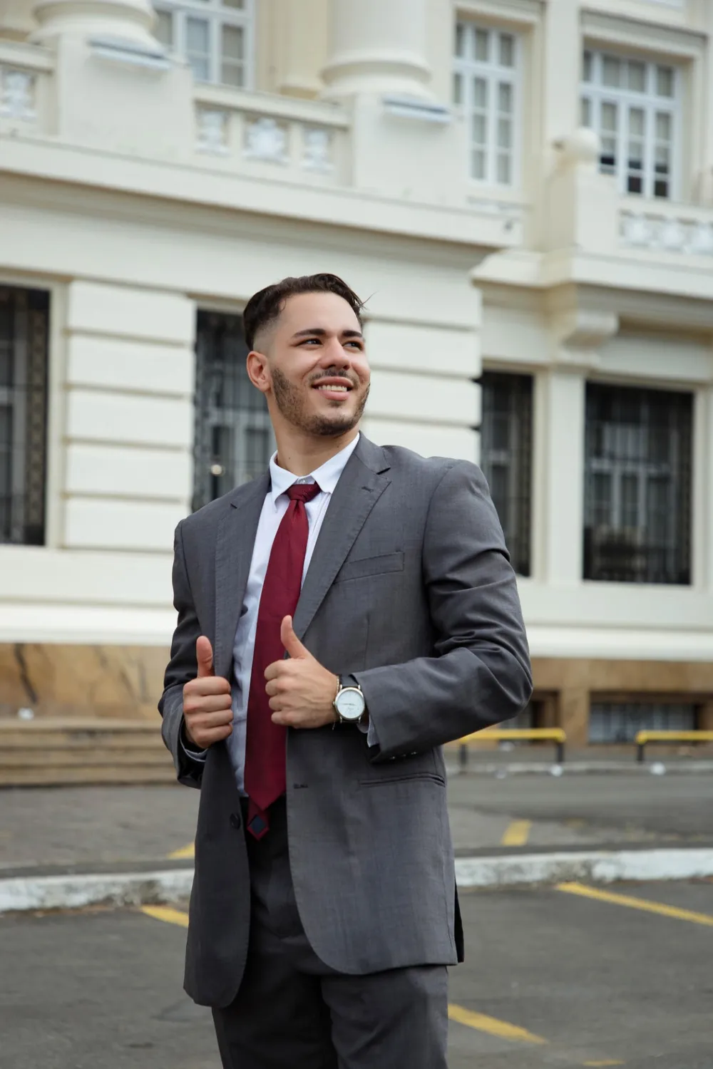 Lucas Tupiná de terno cinza e gravata vinho, sorrindo, em frente a um prédio histórico